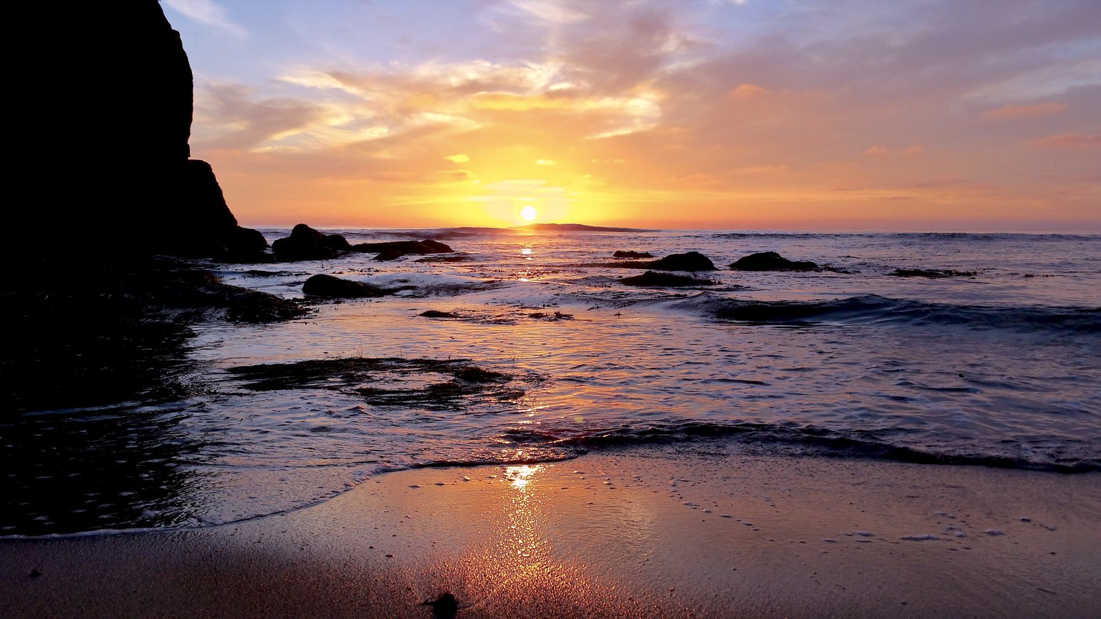 Southern California sunset over the Pacific: a dark sea cliff on the left, the sun sitting just above the horizon, and warm gold light reflecting across the wet sand and shallow surf.