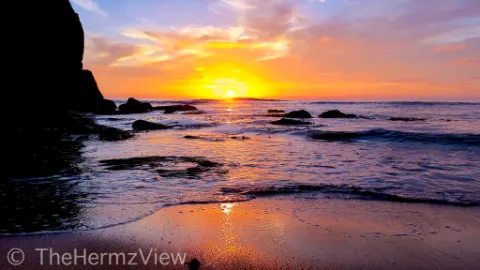 Sunset over a ocean shore with bright golden light reflecting off wet sand, gentle incoming waves, and dark silhouetted coastal rocks.