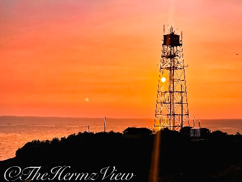 A tall steel lattice tower silhouetted against a deep orange sunset sky over the ocean coastline.
