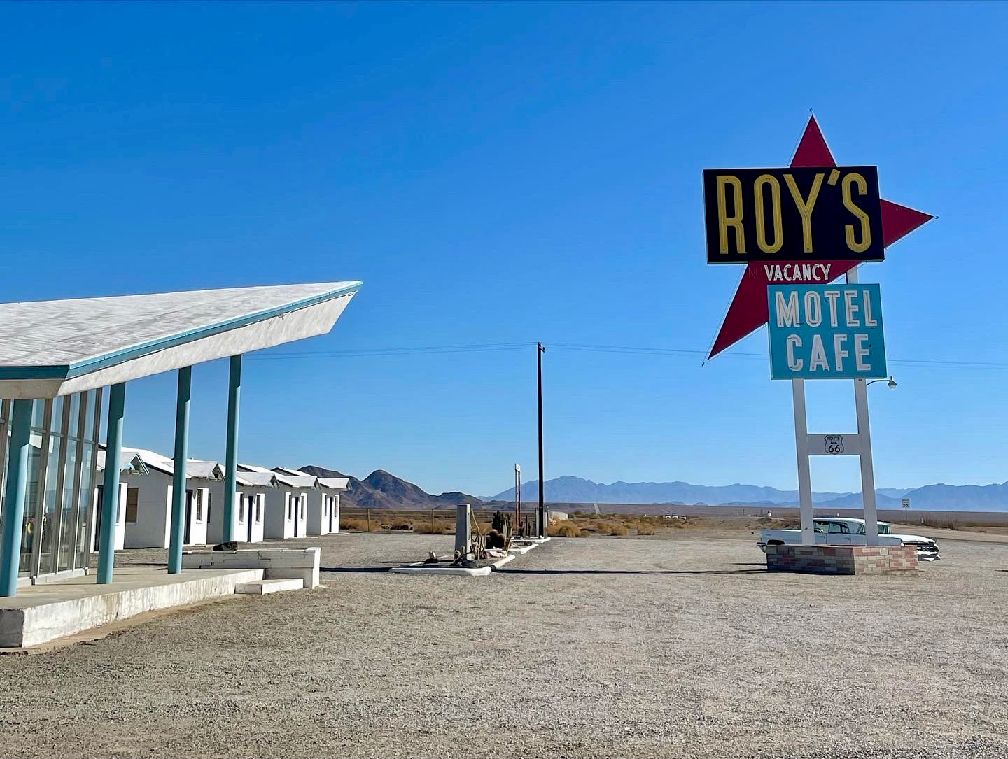 A retro sign reading Roy's Motel Cafe stands in a gravel lot next to a mid-century modern building under a clear blue sky.