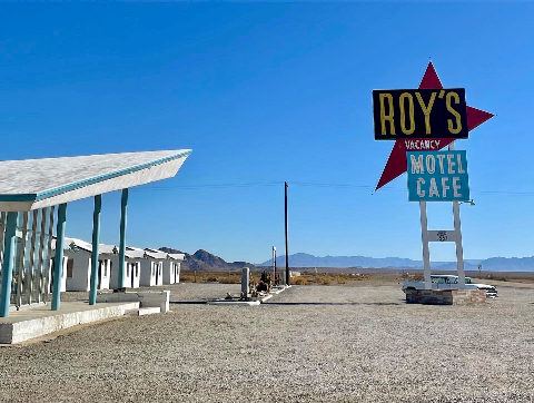 A retro sign reading Roy's Motel Cafe stands in a gravel lot next to a mid-century modern building under a clear blue sky.