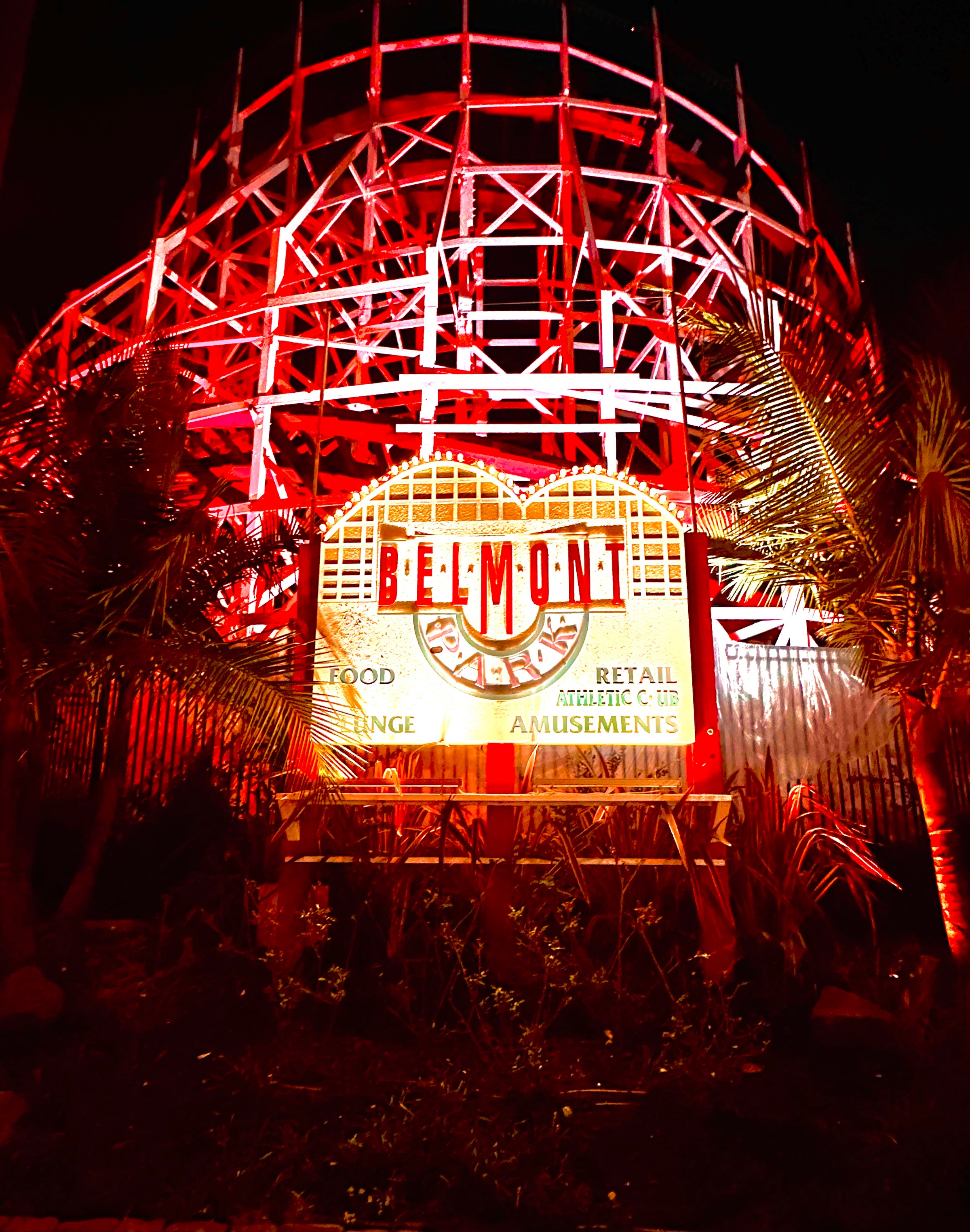 An illuminated vintage sign reading Belmont Park positioned in front of a wooden roller coaster structure glowing in intense red light at night.