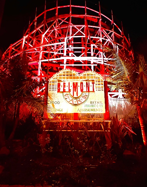 An illuminated vintage sign reading Belmont Park positioned in front of a wooden roller coaster structure glowing in intense red light at night.