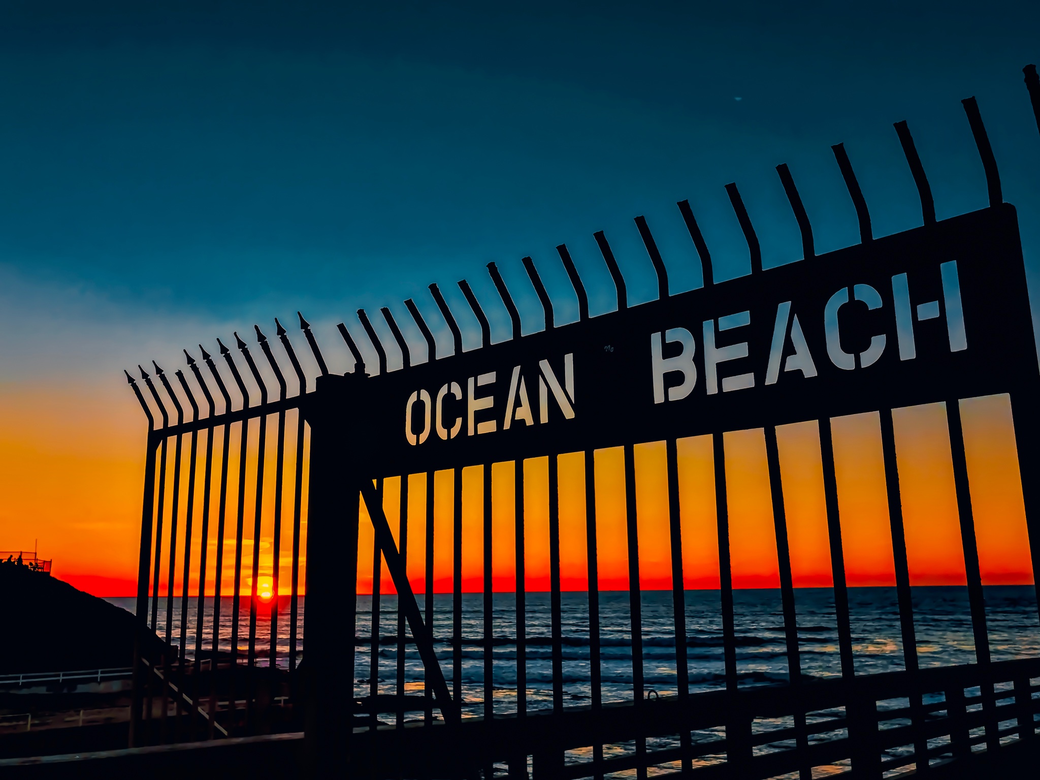 Silhouette of a metal gate with cutout lettering that reads OCEAN BEACH, set against a fiery ocean sunset and deep blue sky.