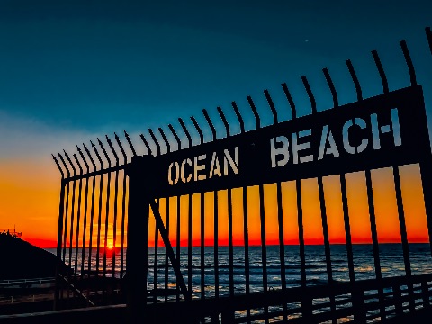 Silhouette of a metal gate with cutout lettering that reads OCEAN BEACH, set against a fiery ocean sunset and deep blue sky.