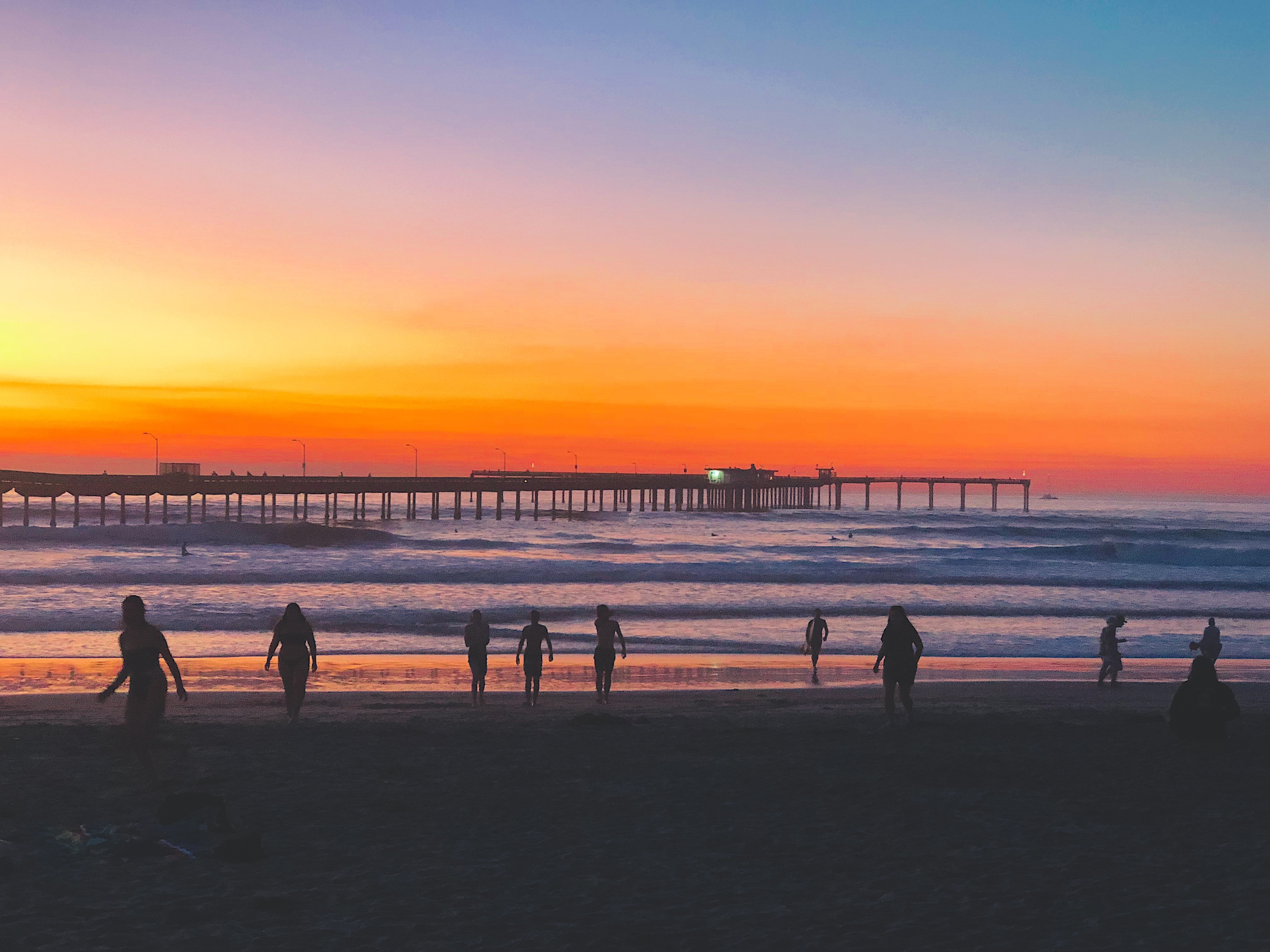 Silhouetted figures walk along a sandy beach under a vibrant orange, magenta, and blue twilight sky with a long pier extending into ocean waves.