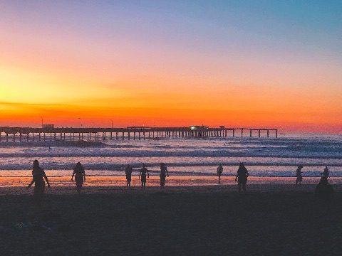 Silhouetted figures walk along a sandy beach under a vibrant orange, magenta, and blue twilight sky with a long pier extending into ocean waves.