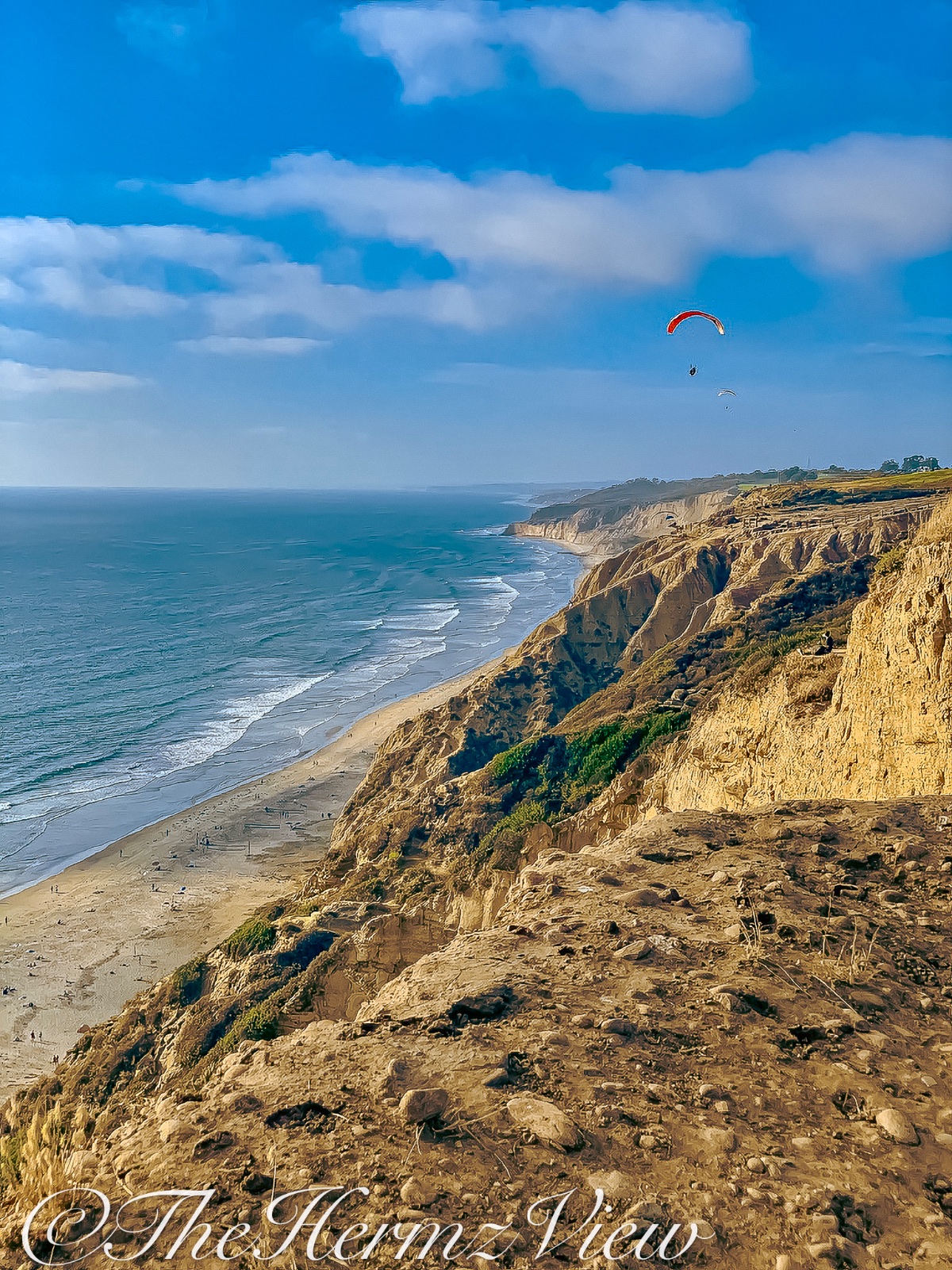 High-angle view of steep coastal cliffs meeting a sandy beach and ocean under a blue sky with a paraglider above.
