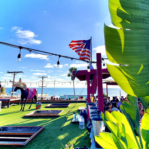 A sunny rooftop patio with green turf, string lights and an American flag overlooks the Pacific, with a horse statue wearing a sombrero and striped blanket on the left and large tropical leaves framing the right.