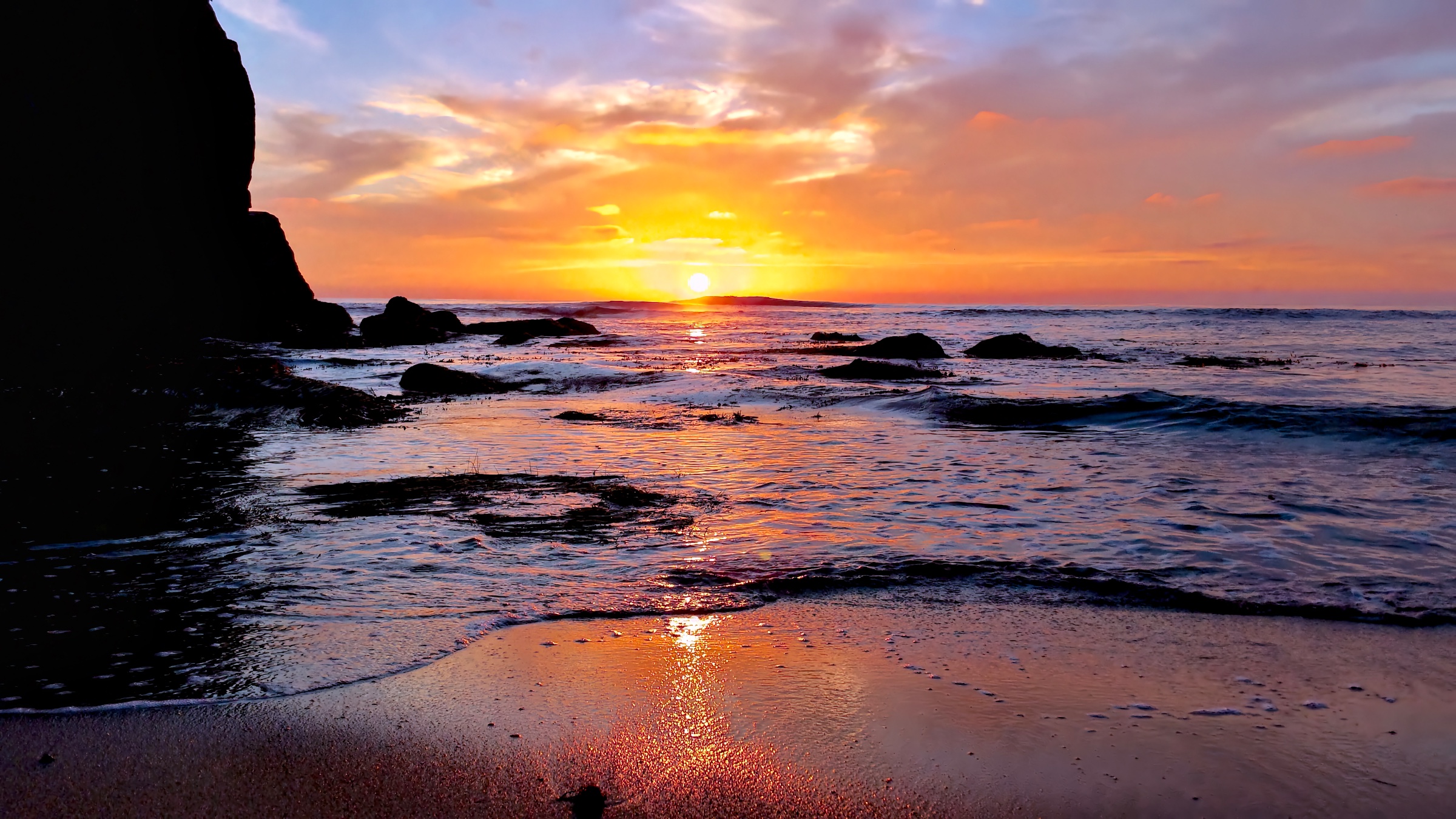 Sunset over a ocean shore with bright golden light reflecting off wet sand, gentle incoming waves, and dark silhouetted coastal rocks.