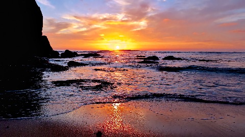 Sunset over a ocean shore with bright golden light reflecting off wet sand, gentle incoming waves, and dark silhouetted coastal rocks.