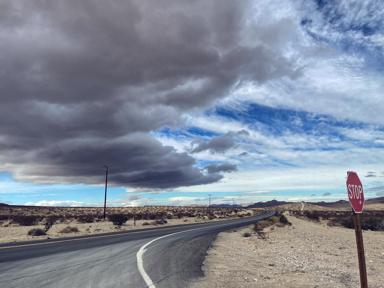 A paved desert highway curves past a stop sign under a dark storm cloud formation and bright blue sky.