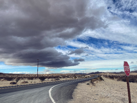 A paved desert highway curves past a stop sign under a dark storm cloud formation and bright blue sky.