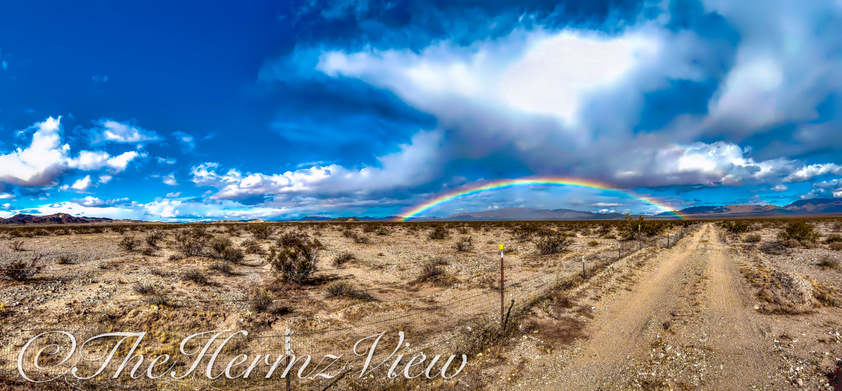 Wide panoramic view of an open desert plain with a dirt road, wire fence line, and a bright rainbow arching in front of distant mountains under a cloudy sky.