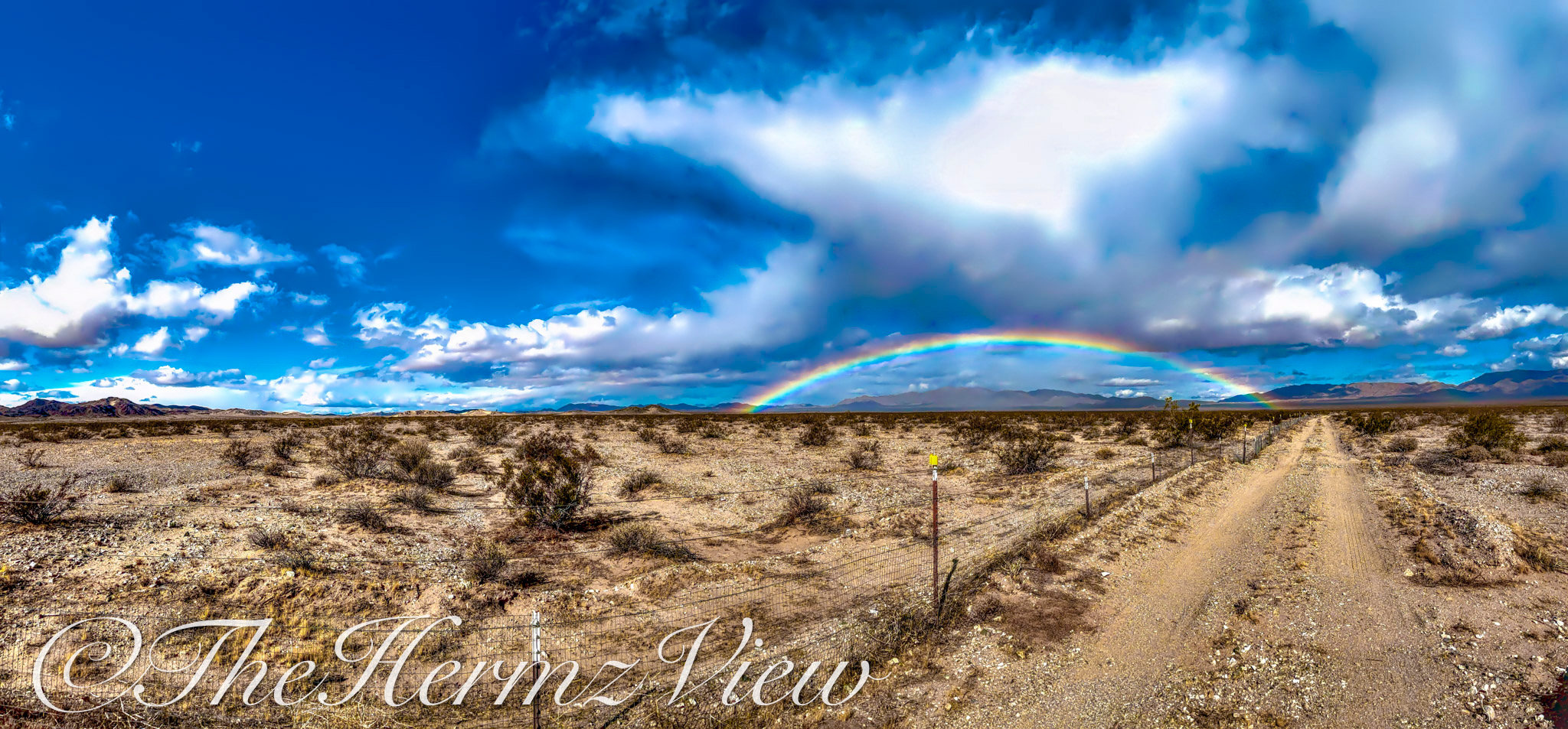 Wide panoramic view of an open desert plain with a dirt road, wire fence line, and a bright rainbow arching in front of distant mountains under a cloudy sky.