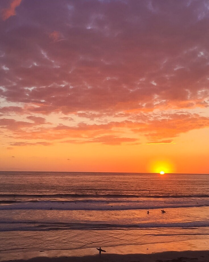 Silhouetted surfers in the ocean and on the shore under a bright orange and purple sunset sky.