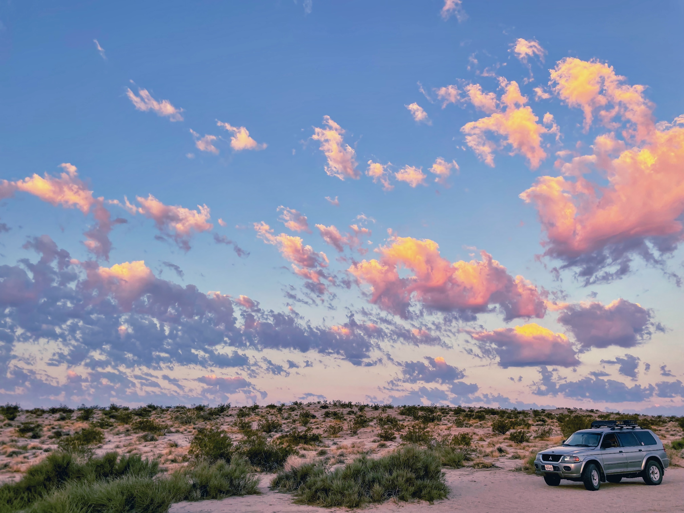 Silver SUV parked on a sandy dirt path among low desert bushes under pink and orange clouds at sunset.