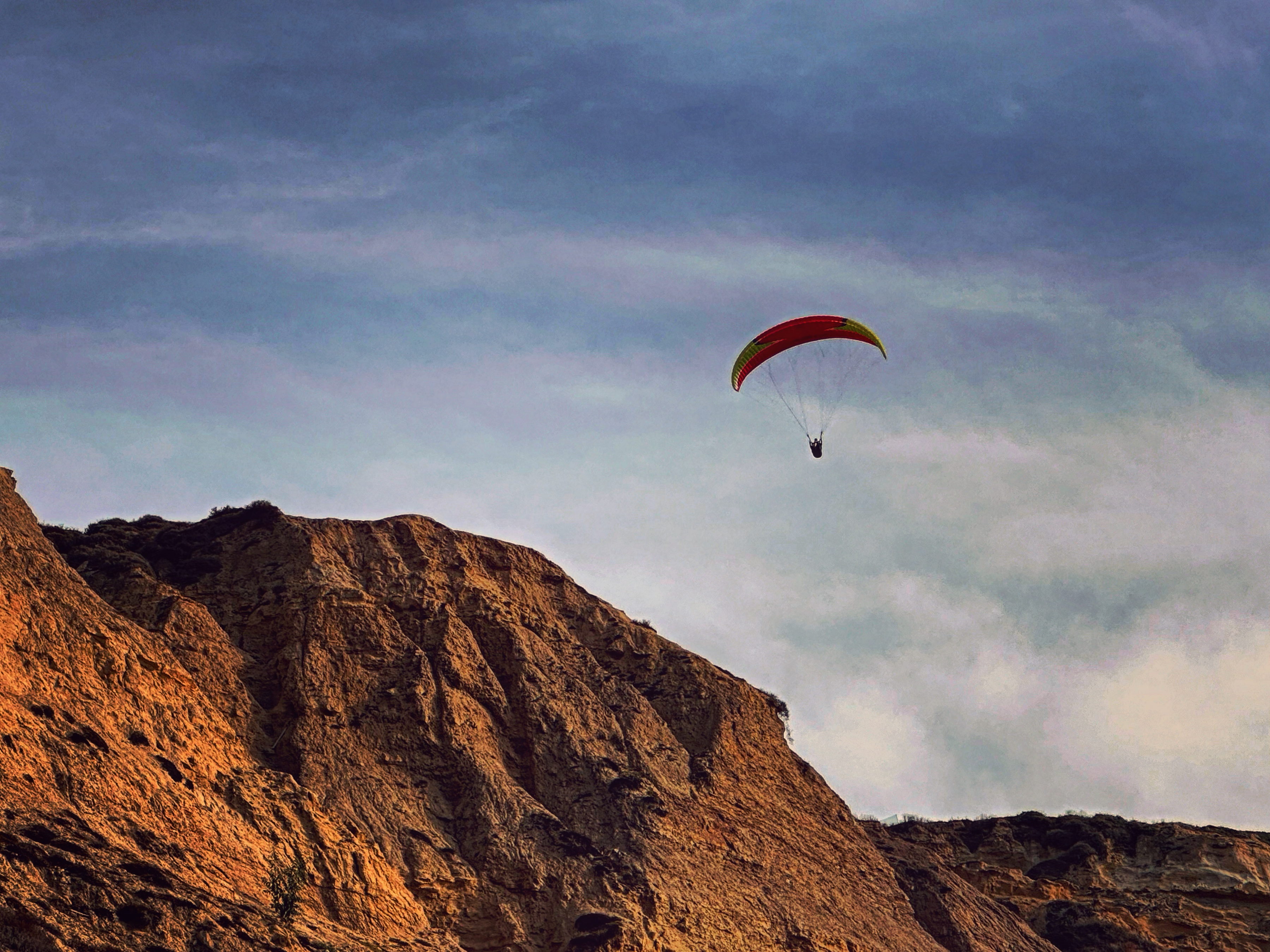 A paraglider with a red and yellow canopy glides through a cloudy blue sky high above steep, sunlit cliffs.