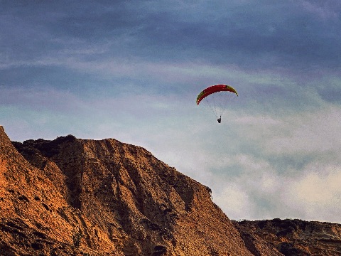 A paraglider with a red and yellow canopy glides through a cloudy blue sky high above steep, sunlit cliffs.