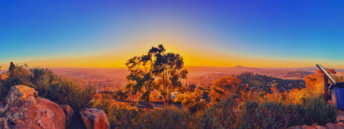 Panoramic sunset view from a rocky hilltop looking out over a wide valley, with a silhouetted tree centered in front of a bright orange horizon.