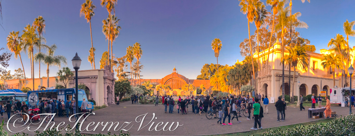A wide panoramic view of a busy public plaza with ornate Spanish-style architecture, tall palm trees, food trucks, and crowds walking under warm golden sunlight.