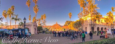 A wide panoramic view of a busy public plaza with ornate Spanish-style architecture, tall palm trees, food trucks, and crowds walking under warm golden sunlight.