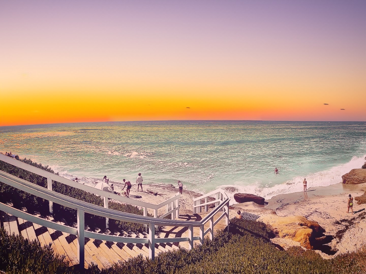 Wooden staircase leading down a vegetation-covered slope toward a rocky cove beach under a warm sunset horizon.