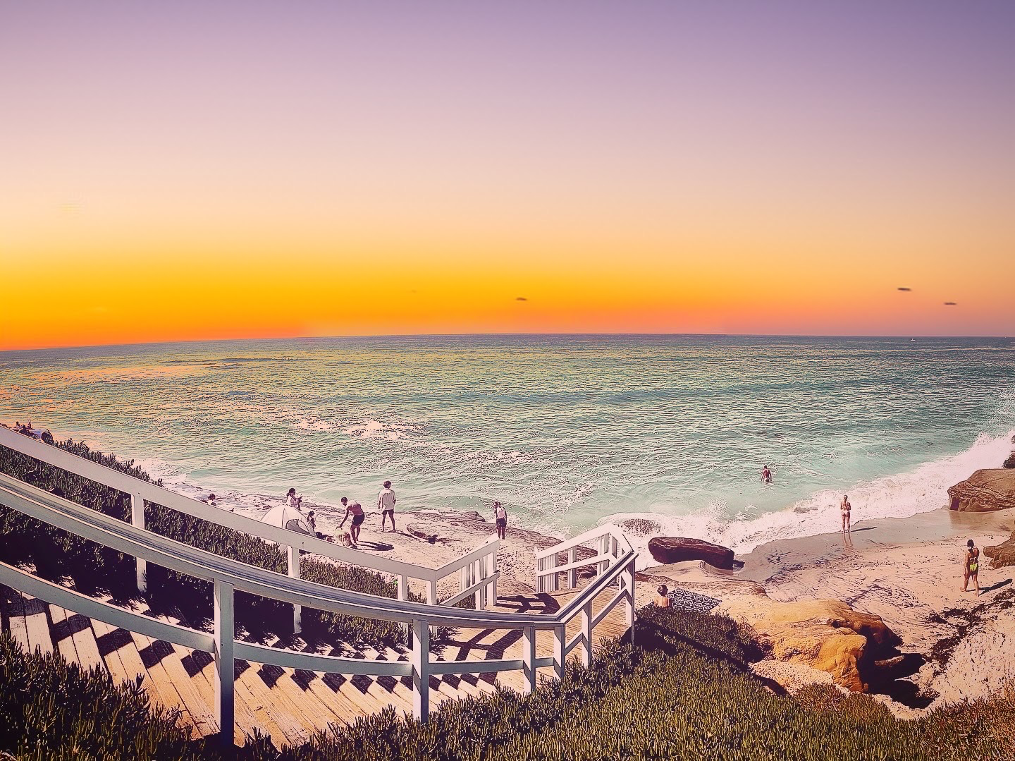 Wooden staircase leading down a vegetation-covered slope toward a rocky cove beach under a warm sunset horizon.