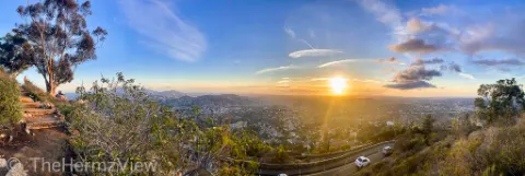 Panoramic sunset view looking down from a hillside path onto a vast valley with a winding road below and bright sun low on the horizon.