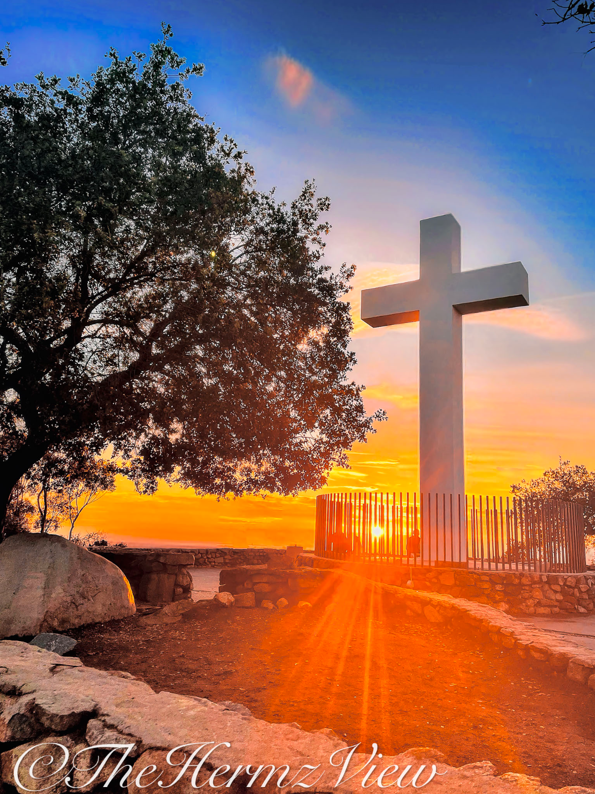 A large white cross stands atop a stone terrace against a bright orange sunset and blue sky, framed by a silhouetted tree on the left.