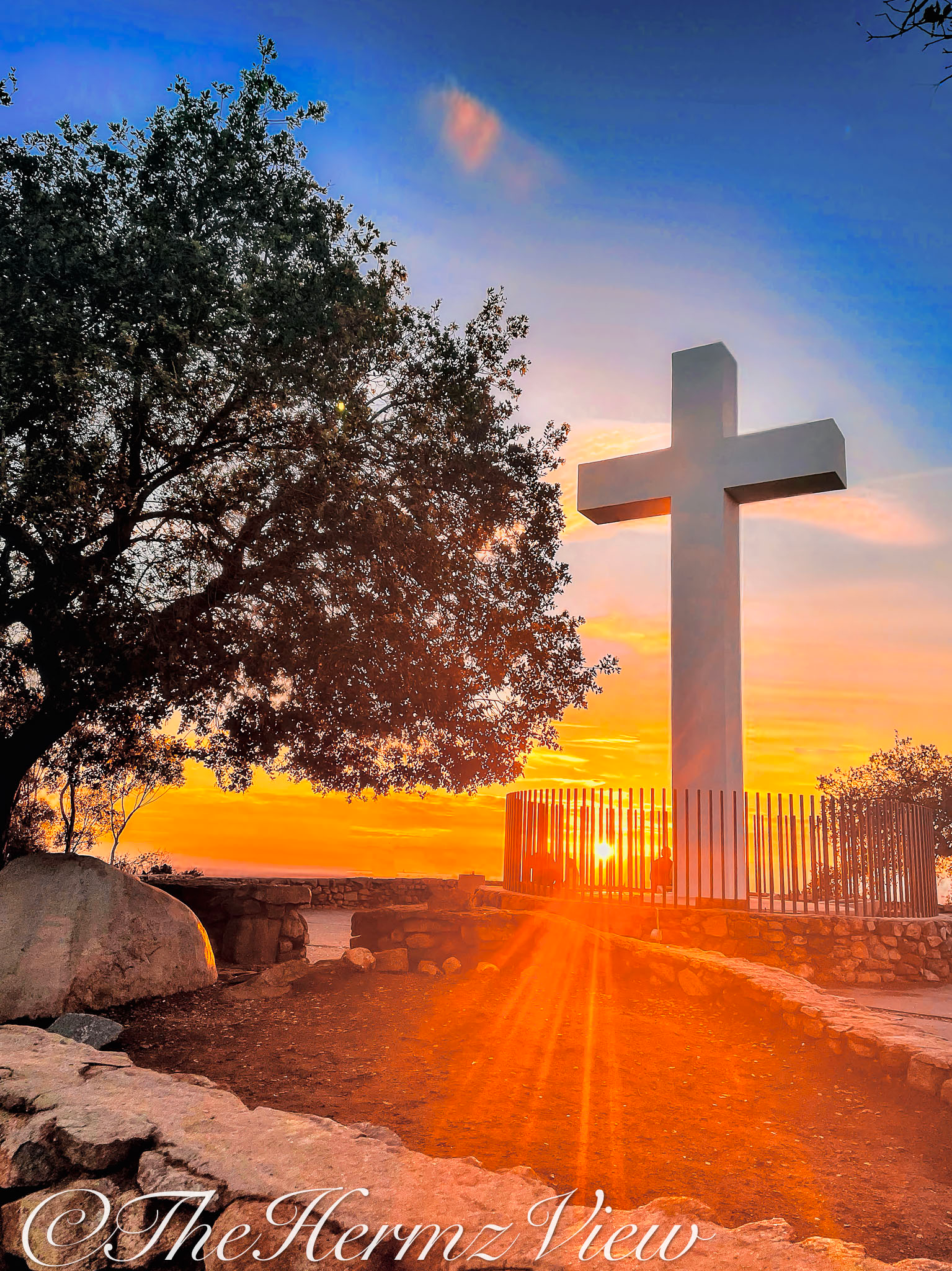 A large white cross stands atop a stone terrace against a bright orange sunset and blue sky, framed by a silhouetted tree on the left.