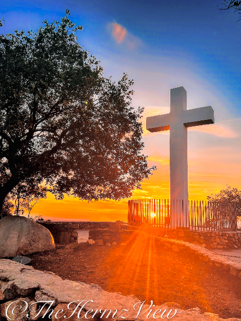 A large white cross stands atop a stone terrace against a bright orange sunset and blue sky, framed by a silhouetted tree on the left.