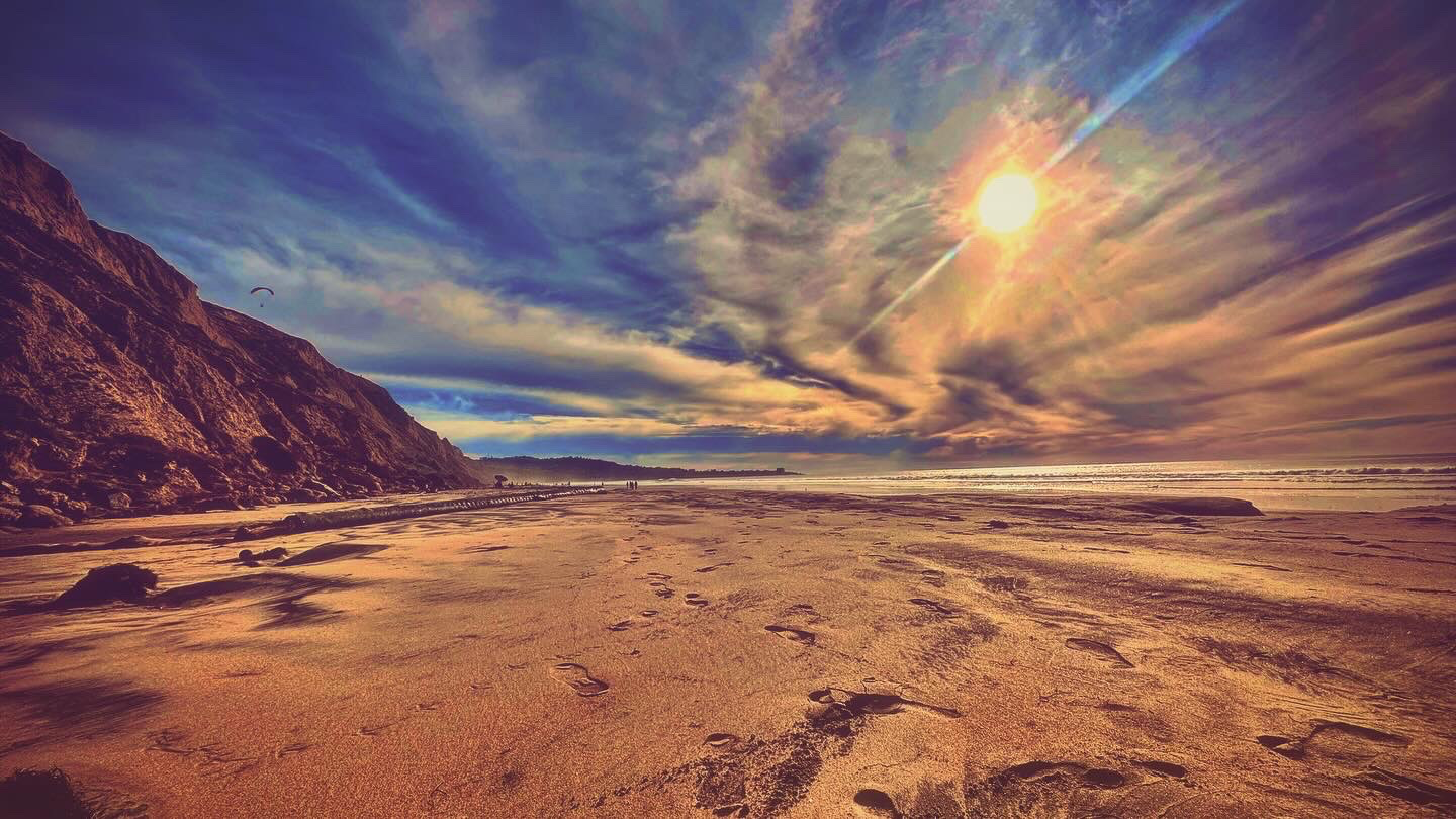 Footprints lead across a wide sandy beach bordered by high coastal cliffs under a bright sun and cloud-streaked sky.