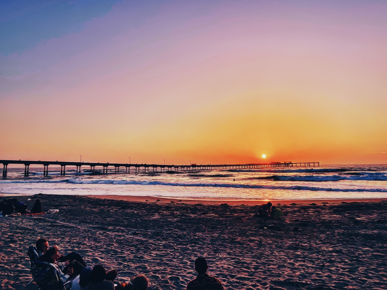 Silhouetted people gathered on a sandy beach watching the sun set on the ocean horizon beside a long pier.