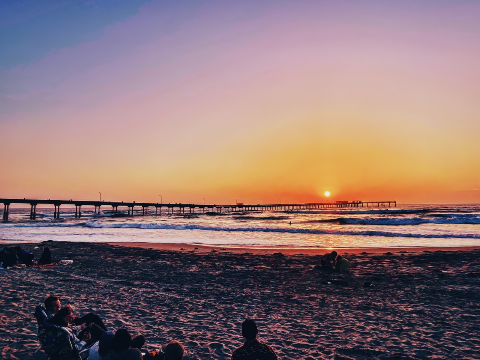 Silhouetted people gathered on a sandy beach watching the sun set on the ocean horizon beside a long pier.