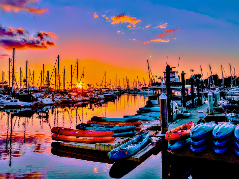 Colorful kayaks lined up on a floating dock at a marina under a bright orange and purple sunset sky.
