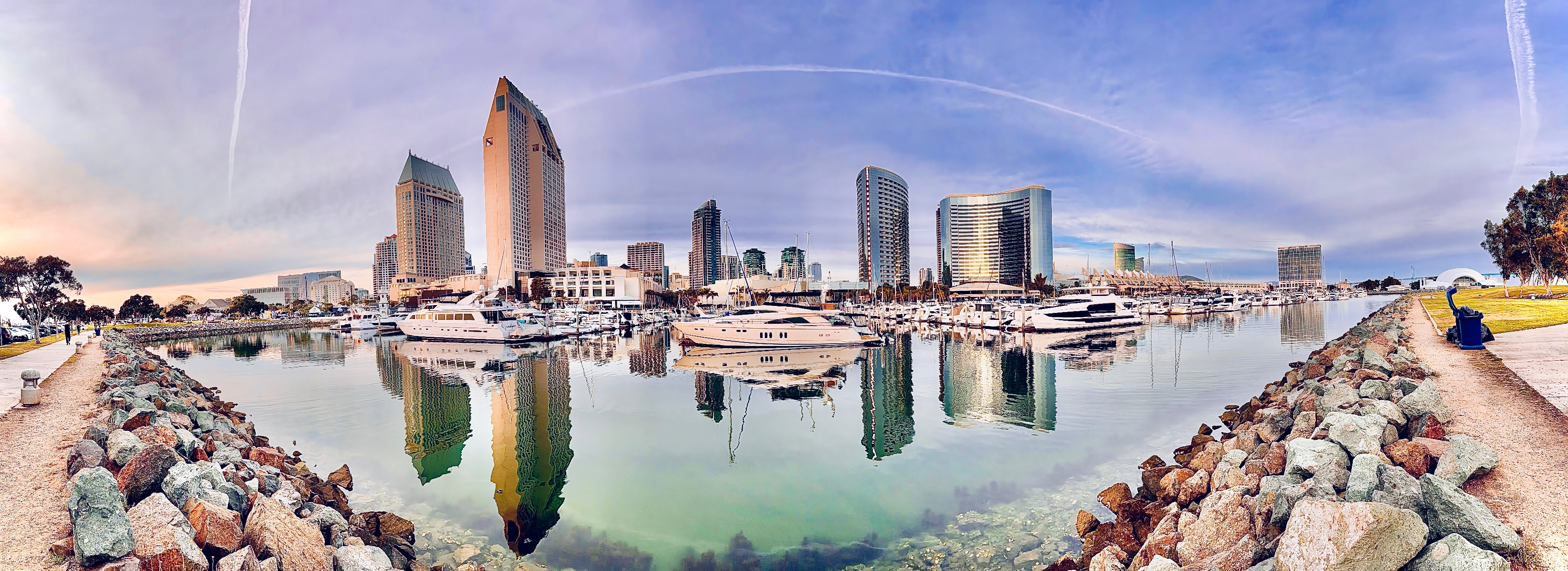 Wide panoramic view of high-rise city buildings and docked yachts reflecting off still harbor waters, framed by rocky shorelines on both sides.