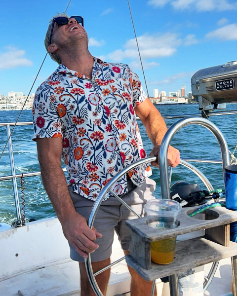 Hermz wearing sunglasses and a patterned shirt stands at the stainless steel wheel of a sailboat on open water with a city skyline in the background.
