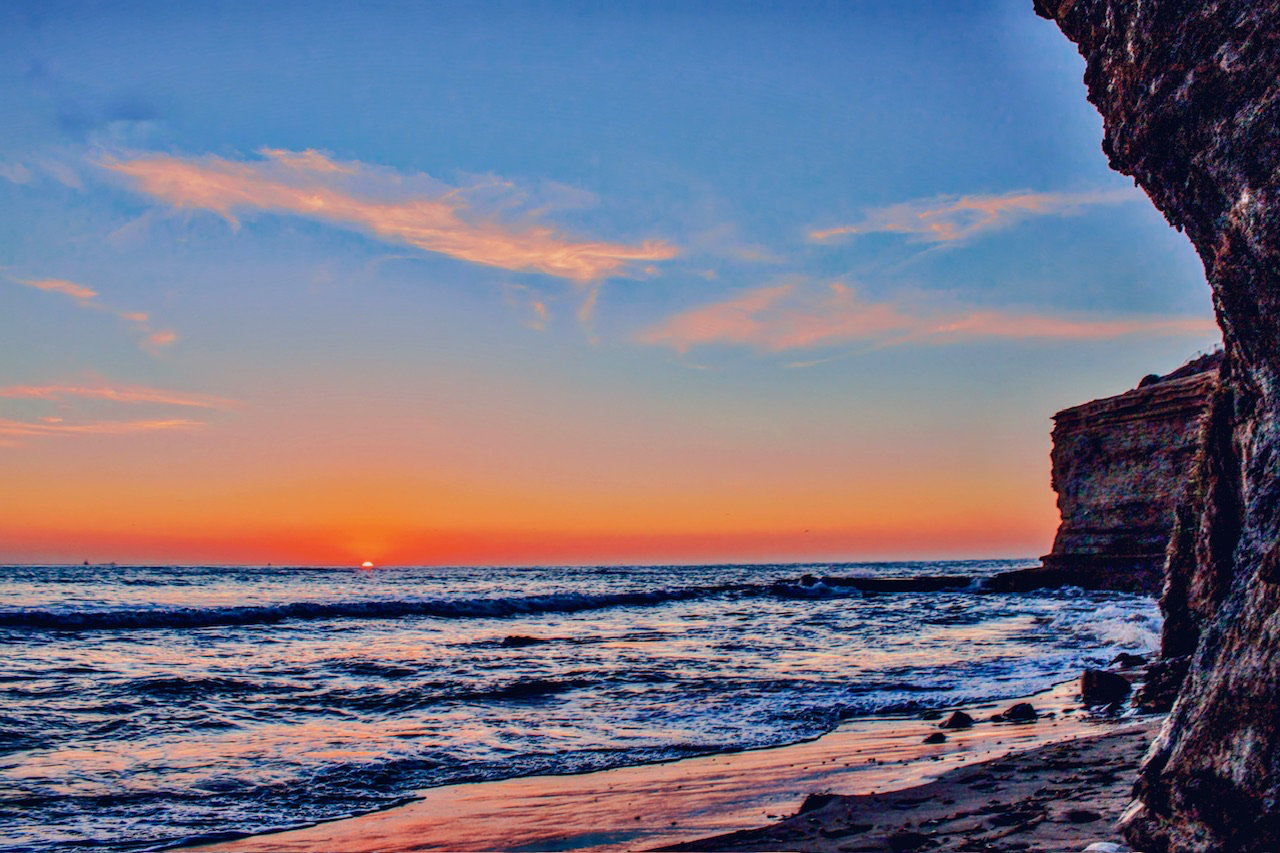 A sunset ocean scene framed by dark rocky cliffs on the right, with gentle waves brushing the shoreline under a blue and orange sky.