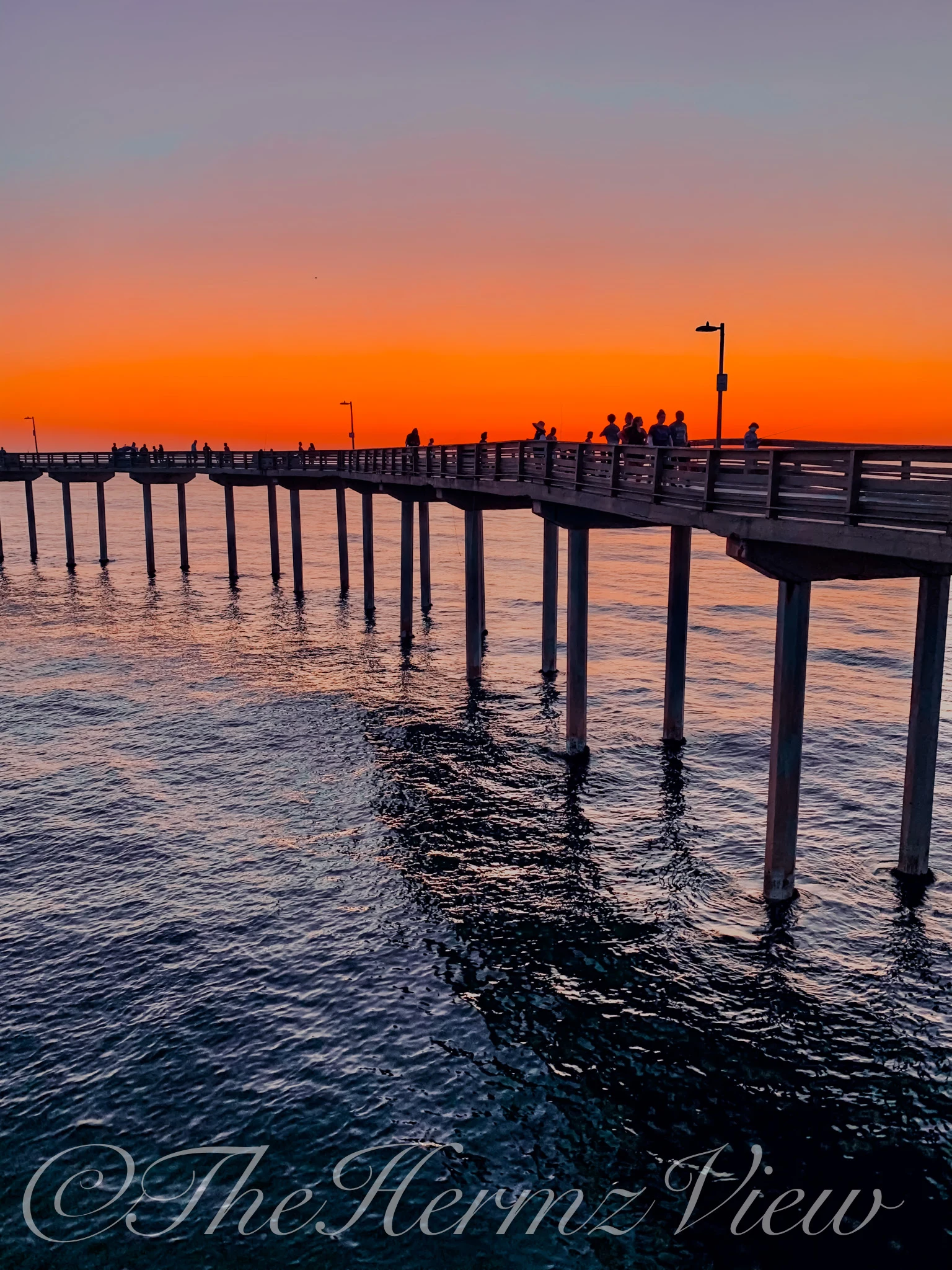 A long ocean pier silhouetted against a brilliant orange and purple sunset sky as dark waters ripple below.