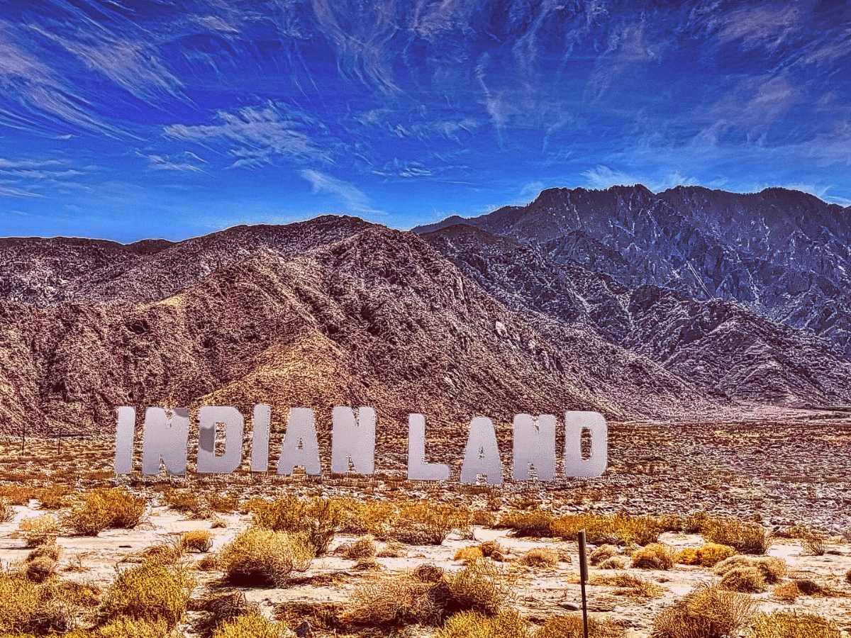 Large white capital letters spelling INDIAN LAND standing in a dry desert landscape against rugged mountains under a blue sky.
