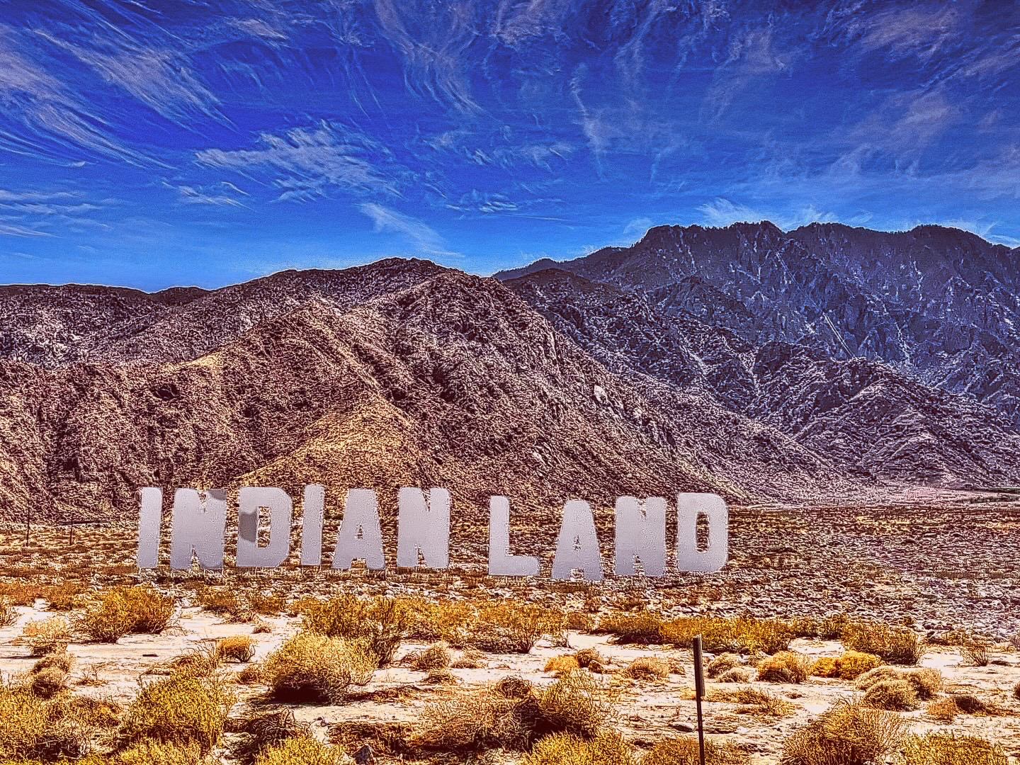 Large white capital letters spelling INDIAN LAND standing in a dry desert landscape against rugged mountains under a blue sky.