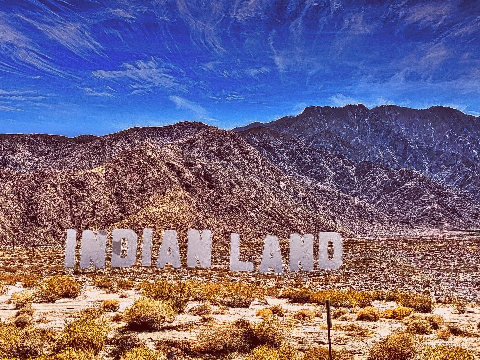 Large white capital letters spelling INDIAN LAND standing in a dry desert landscape against rugged mountains under a blue sky.