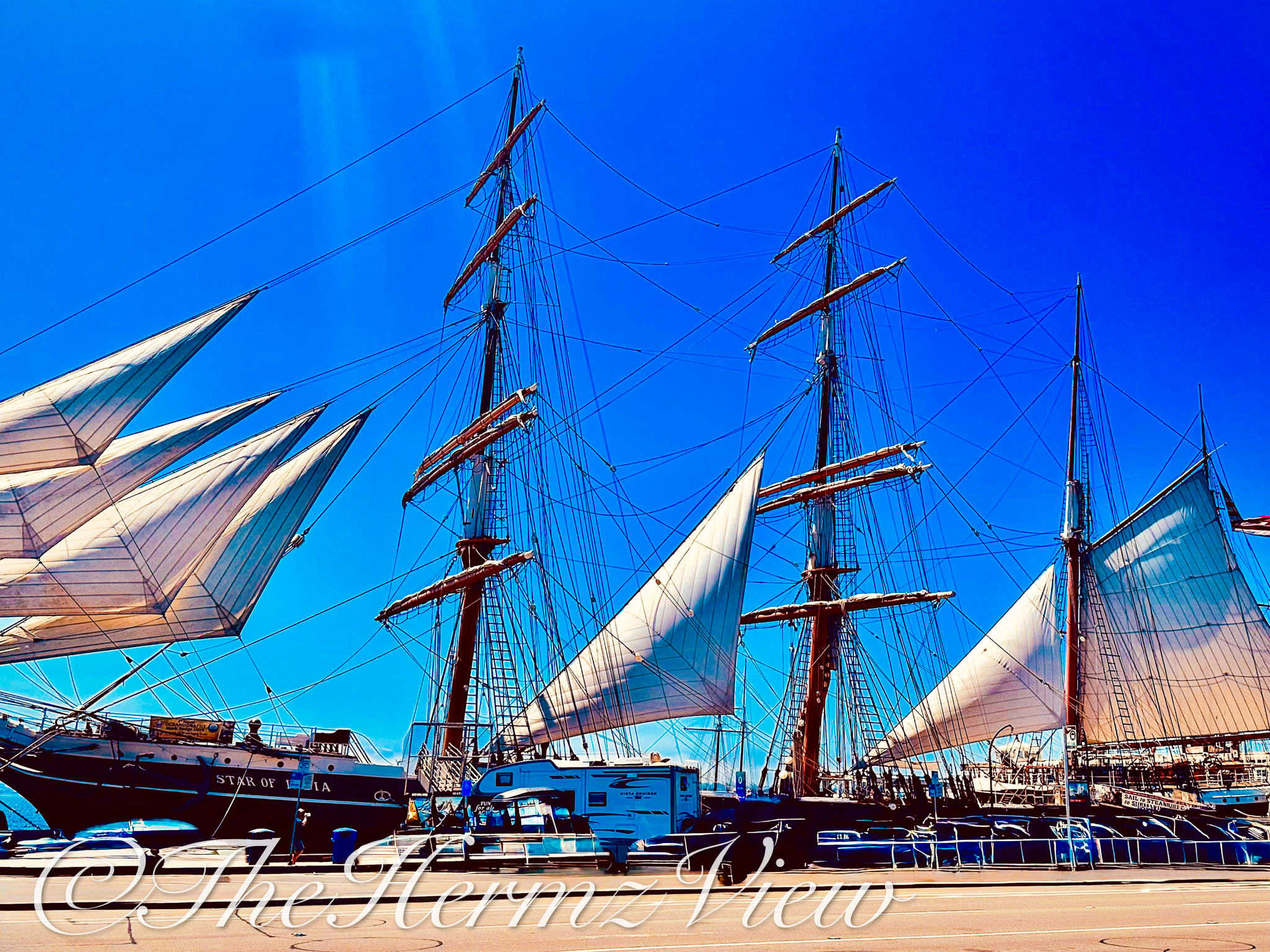 A historic tall ship with white sails and intricate rigging moored at a harbor waterfront under a clear blue sky.
