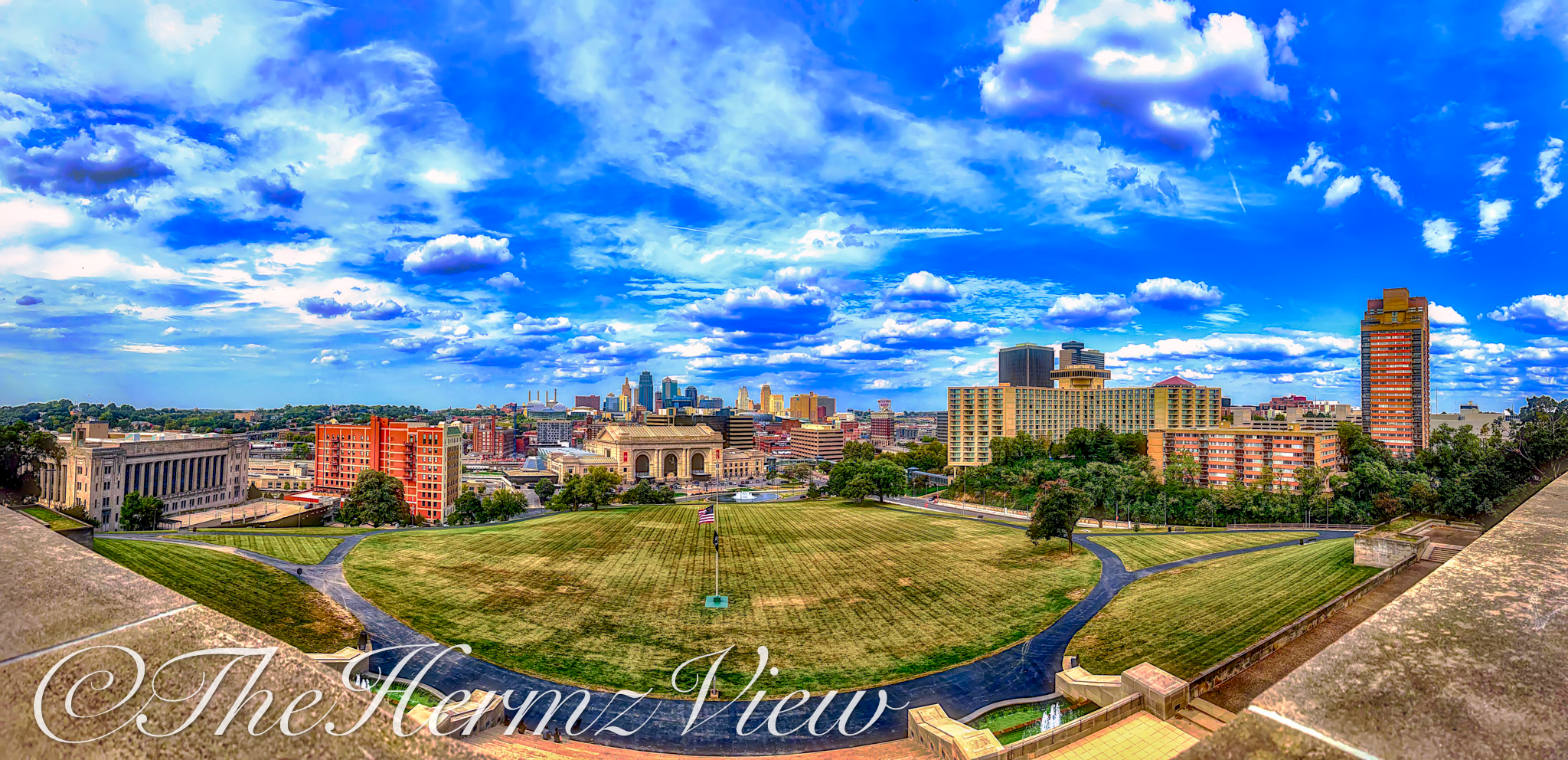 High-angle wide panoramic view of an expansive green lawn, flagpole, city skyline, and bright blue sky with white clouds framed by a stone terrace ledge.
