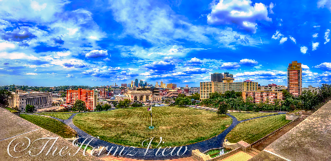 High-angle wide panoramic view of an expansive green lawn, flagpole, city skyline, and bright blue sky with white clouds framed by a stone terrace ledge.