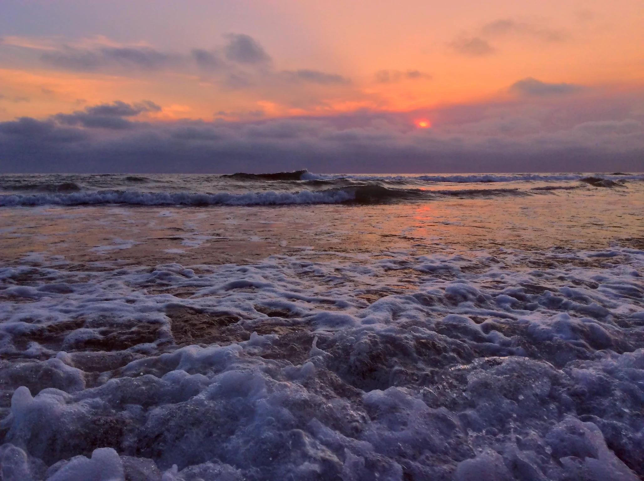 White foamy ocean wave washing onto the shore at sunset under a sky filled with orange and lavender clouds.