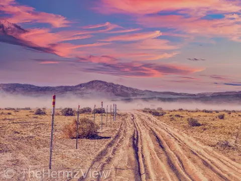 A heavily rutted dirt road in an arid desert landscape stretches toward distant mountains under a vivid pink and purple sunset sky.