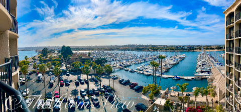 High-angle panoramic view looking out across a marina filled with boats, framed by hotel balconies and palm trees beneath a blue sky with wispy clouds.