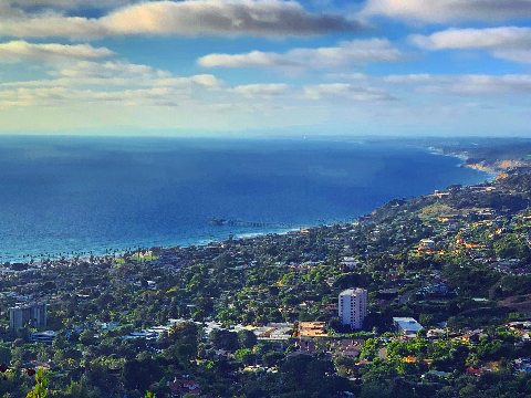 Elevated view overlooking a lush coastal town with a wooden pier extending into the blue ocean under scattered clouds.
